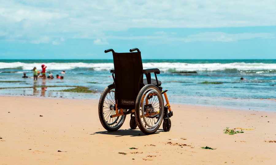 A wheelchair on the sand at the water's edge, with the sea and swimmers behind